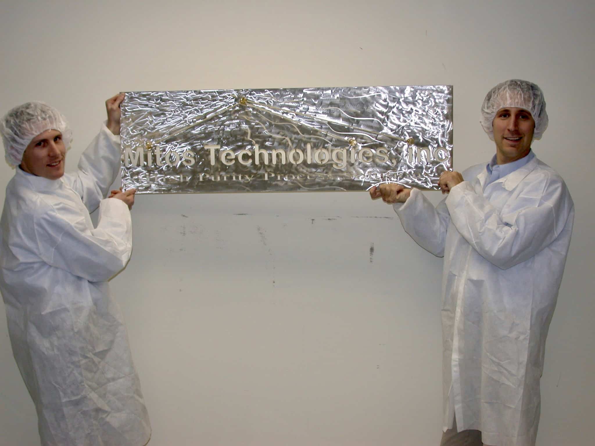 Two people in cleanroom suits holding a Mitos Technologies banner