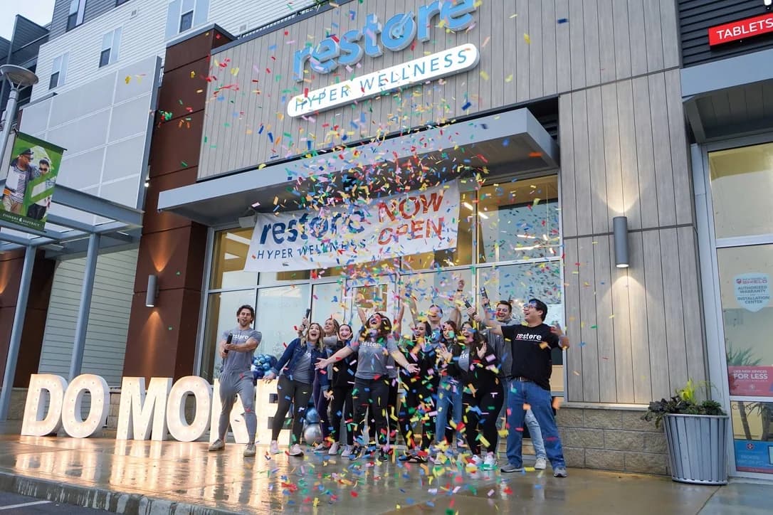 The Restore Hyper Wellness team celebrating a studio opening in a burst of confetti under a NOW OPEN banner