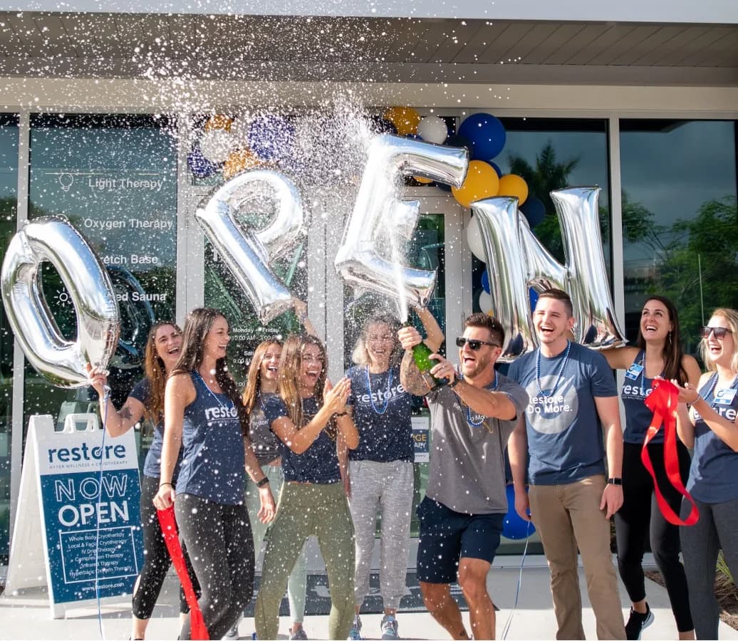 The Restore Hyper Wellness team celebrating a studio opening with champagne under silver OPEN balloons